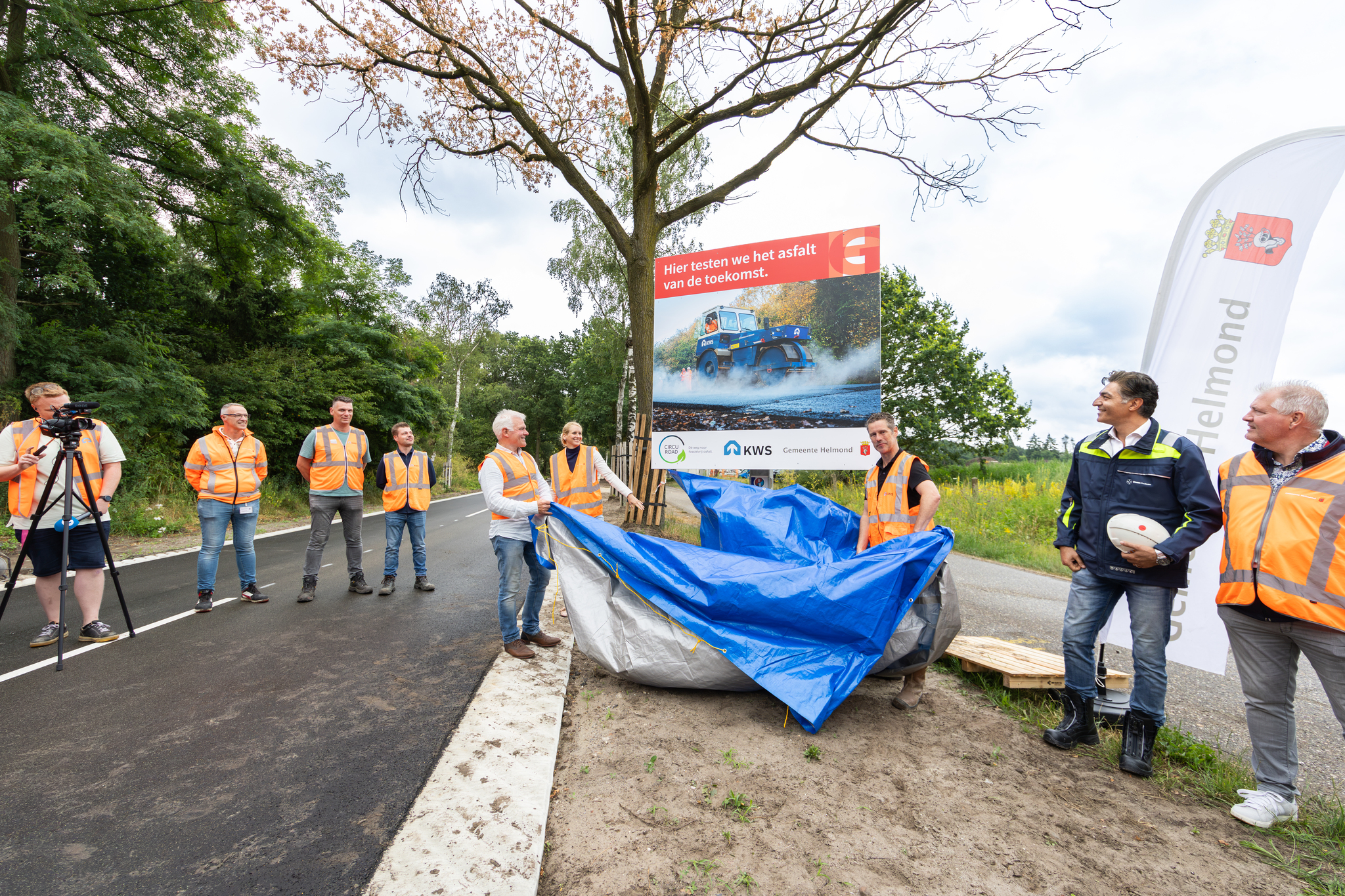 Bord wordt onthuld wat is geplaatst op de weg. Ernaast staan verschillende mensen in een oranje hesje. 